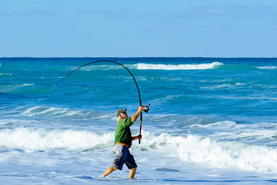 An angler in the surf leaning into a long cast with a bent rod on a bright blue day